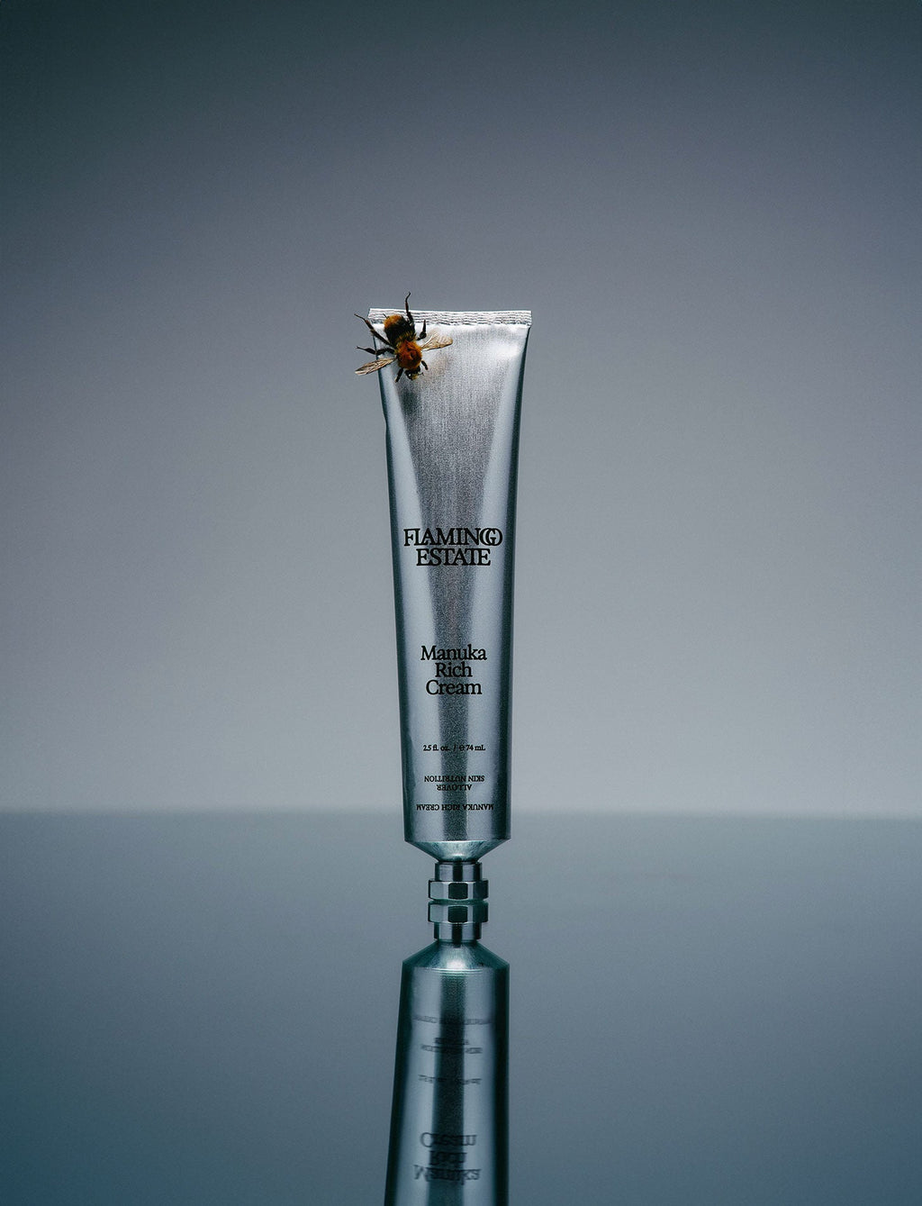 A tube of Flamingo Estate Manuka Rich Cream with a whipped texture, displayed against a grey background. A honey bee rests on the top of the tube.