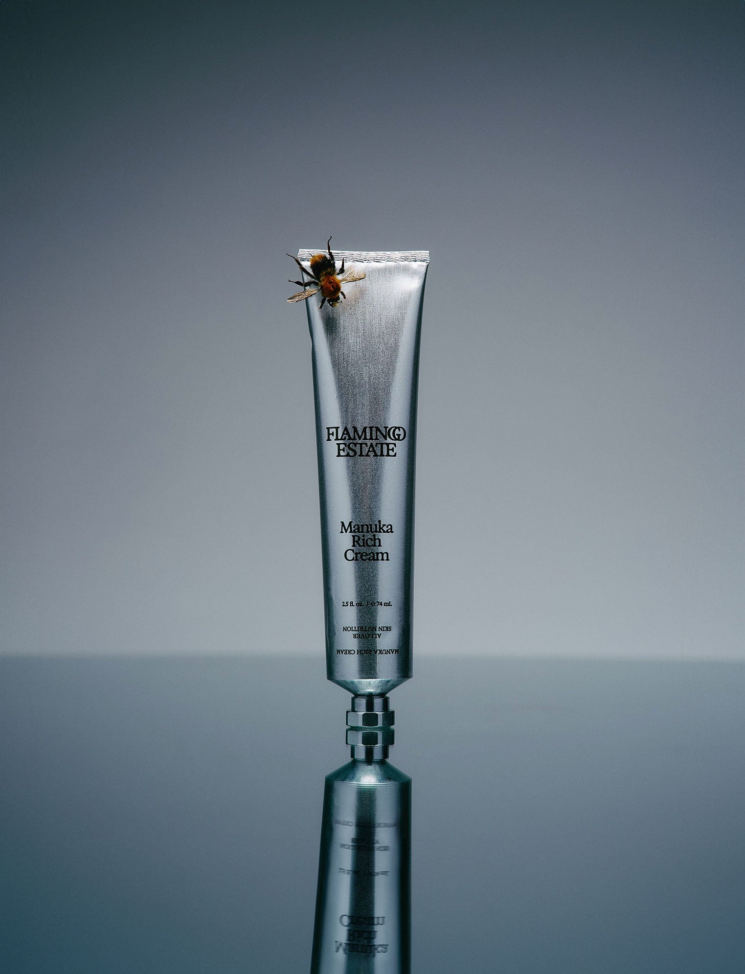 A tube of Flamingo Estate Manuka Rich Cream with a whipped texture, displayed against a grey background. A honey bee rests on the top of the tube.