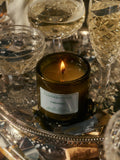 A lit candle with a label that reads 'CORIANDOLI' on a glass surface reflecting the flame, surrounded by champagne glasses and cutlery.
