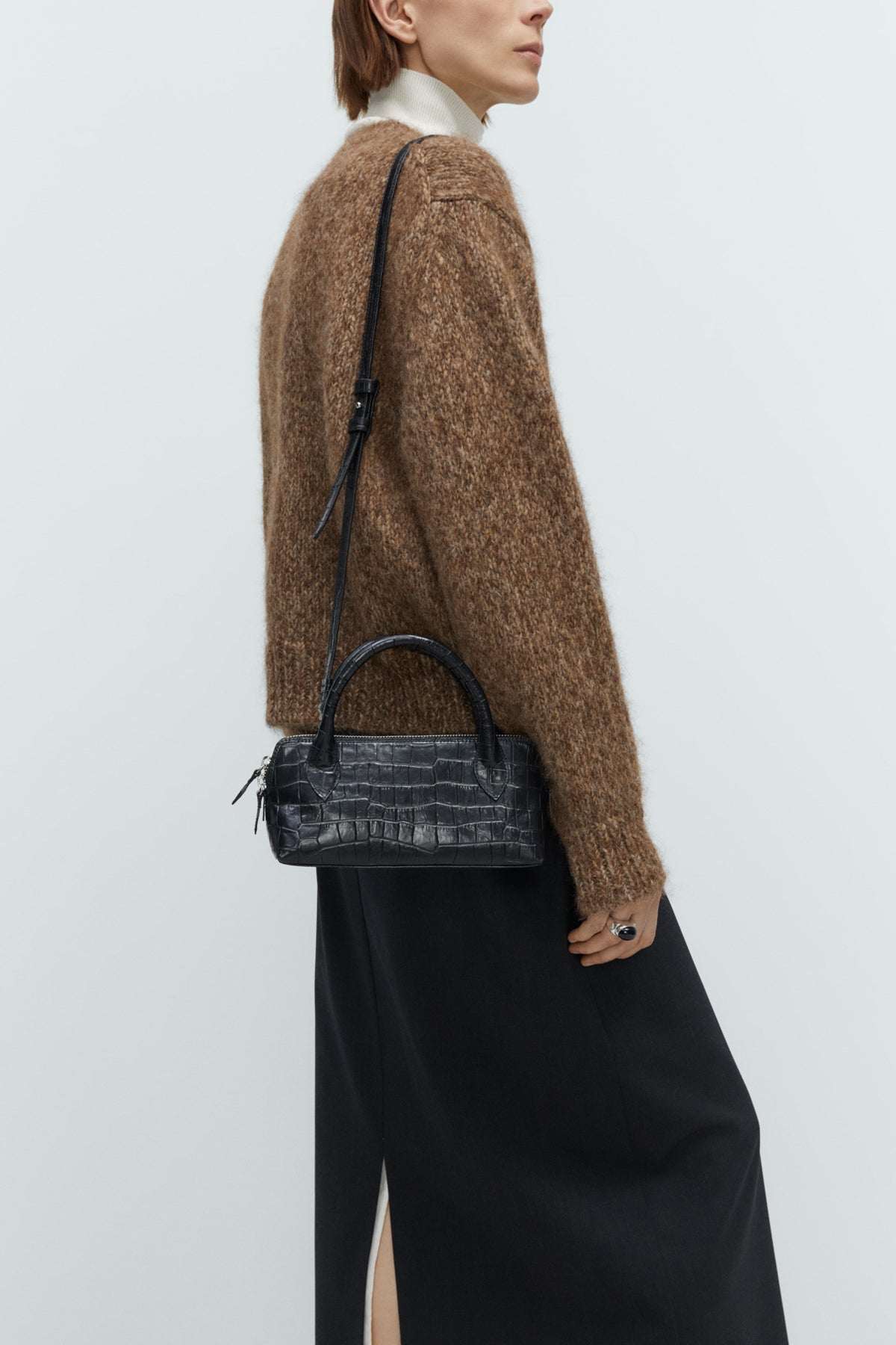 Person wearing a brown sweater and black pants with a black handbag on a white background