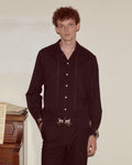 Man wearing a dark brown shirt and pants standing against a plain wall.