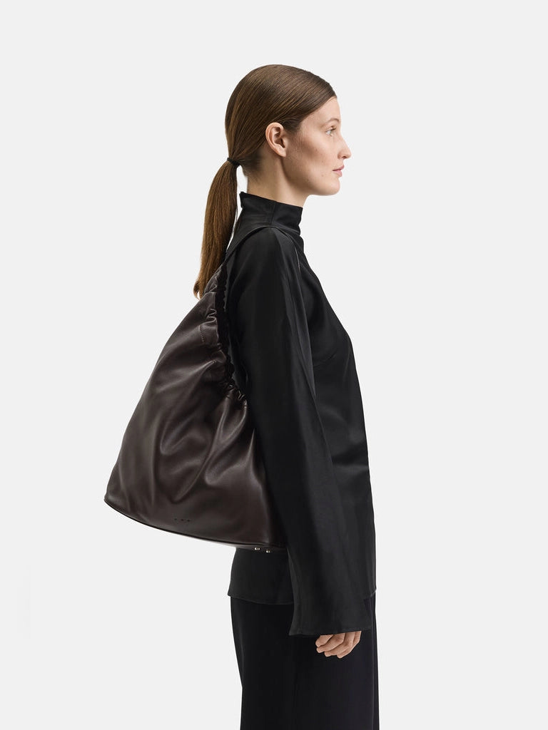 Model carrying Aesther Ekme Demi Lune Cloud in tabacco calfskin with an asymmetrical shape, ruched opening and long shoulder strap.