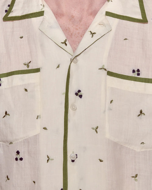 Close-up of a white shirt with green trim and small embroidered details on a pink background