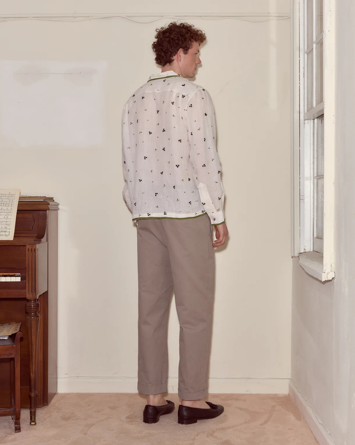 Person wearing a patterned shirt and beige pants standing in a room with a piano and window.