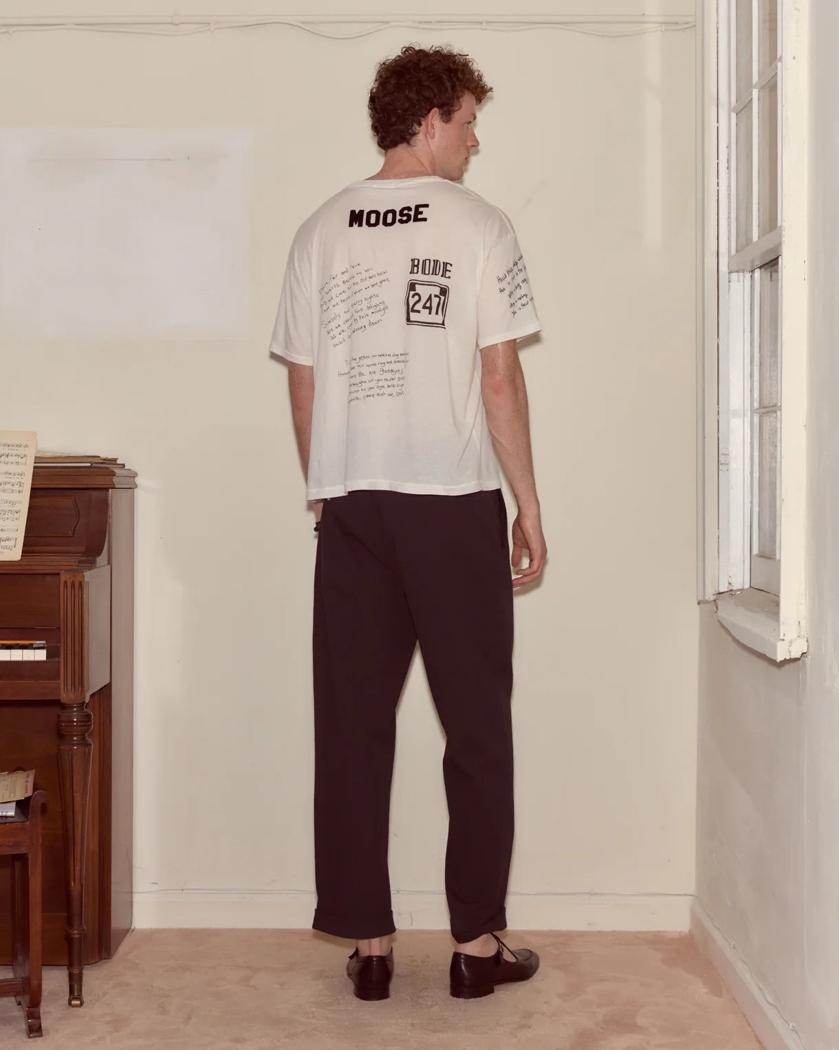 Person wearing a white t-shirt with text, standing in a room with a piano.