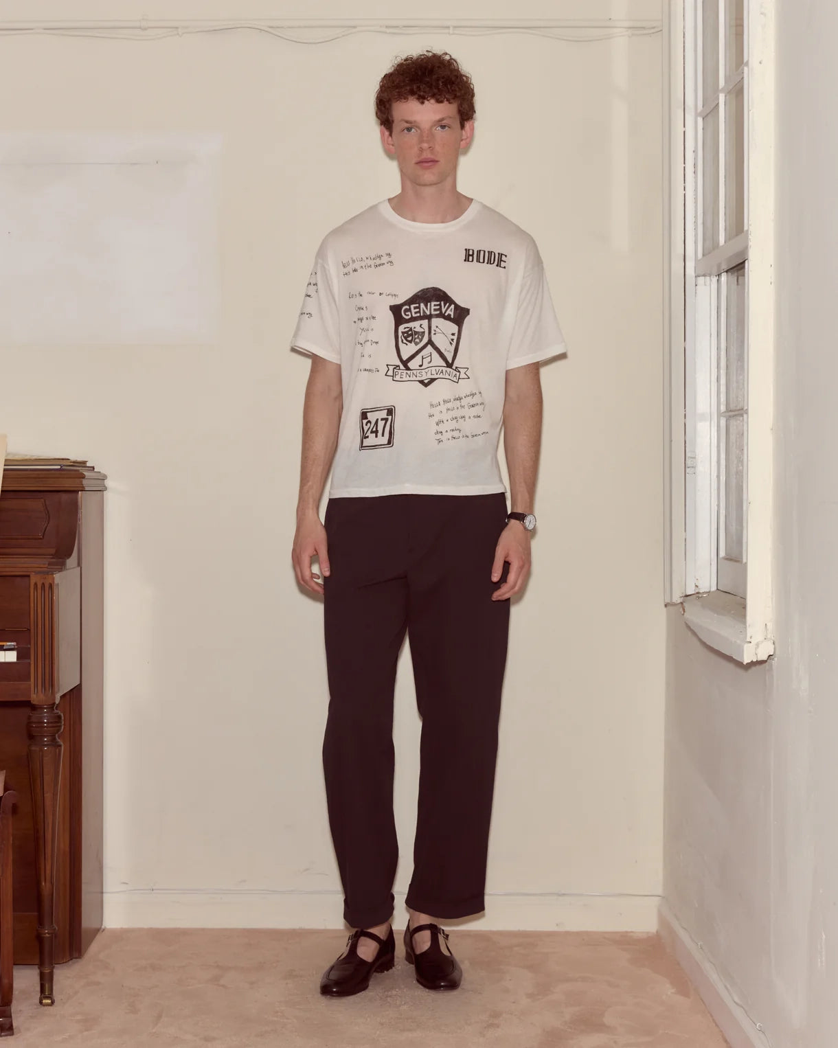 Man wearing a white t-shirt with text and graphics, dark pants, and black shoes in a room.