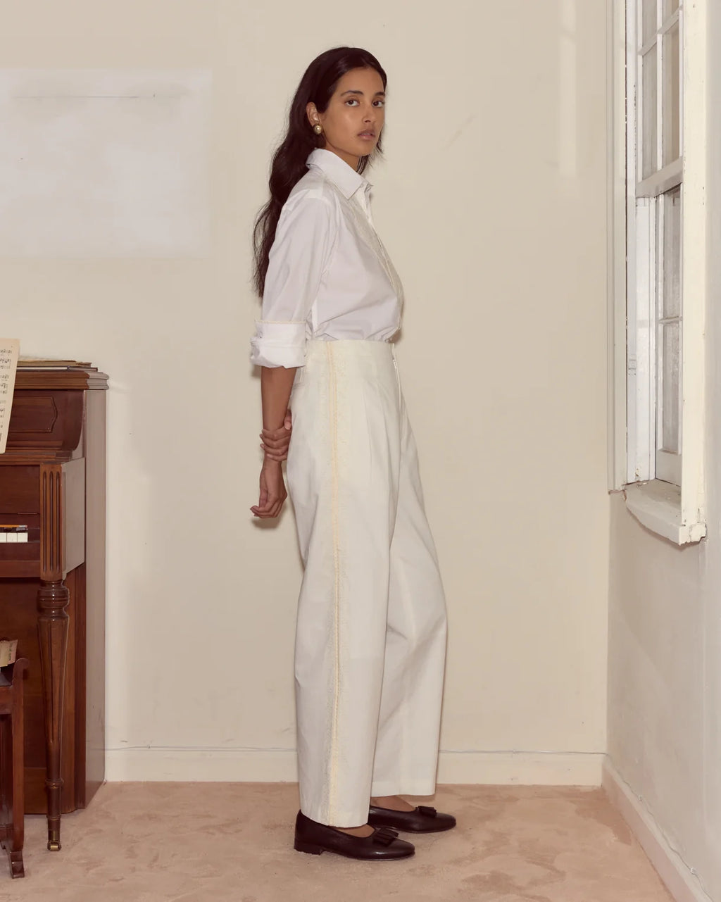 Woman wearing a white shirt and light-colored pants standing in a room with a window and wooden furniture.