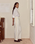 Woman wearing a white shirt and light-colored pants standing in a room with a window and wooden furniture.
