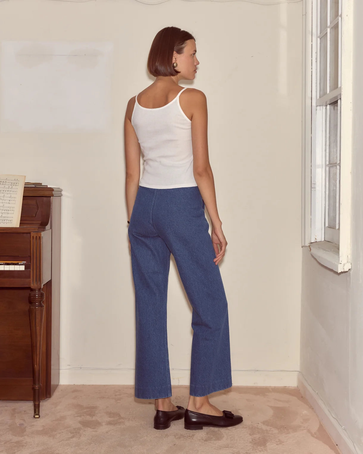 Woman wearing a white tank top and blue jeans standing in a room with a wooden cabinet and window.