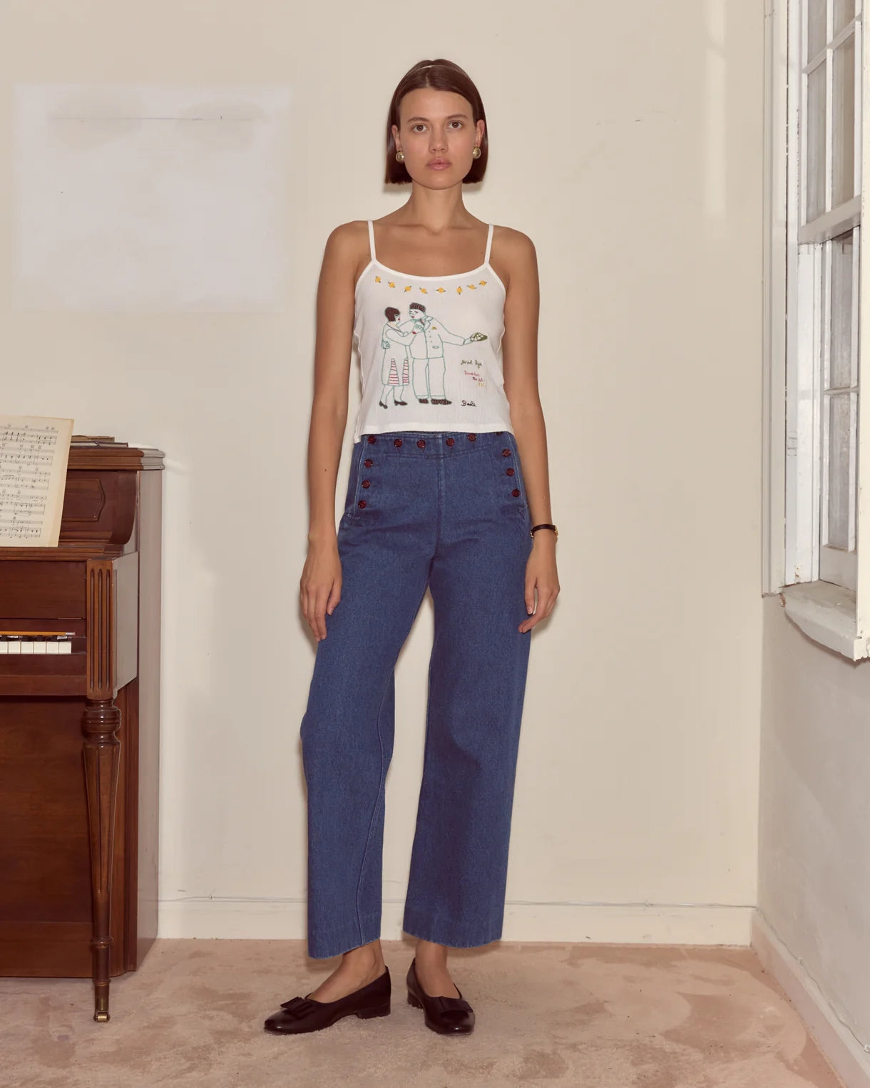Woman wearing a white tank top with a graphic design and blue jeans standing in a room with a piano.
