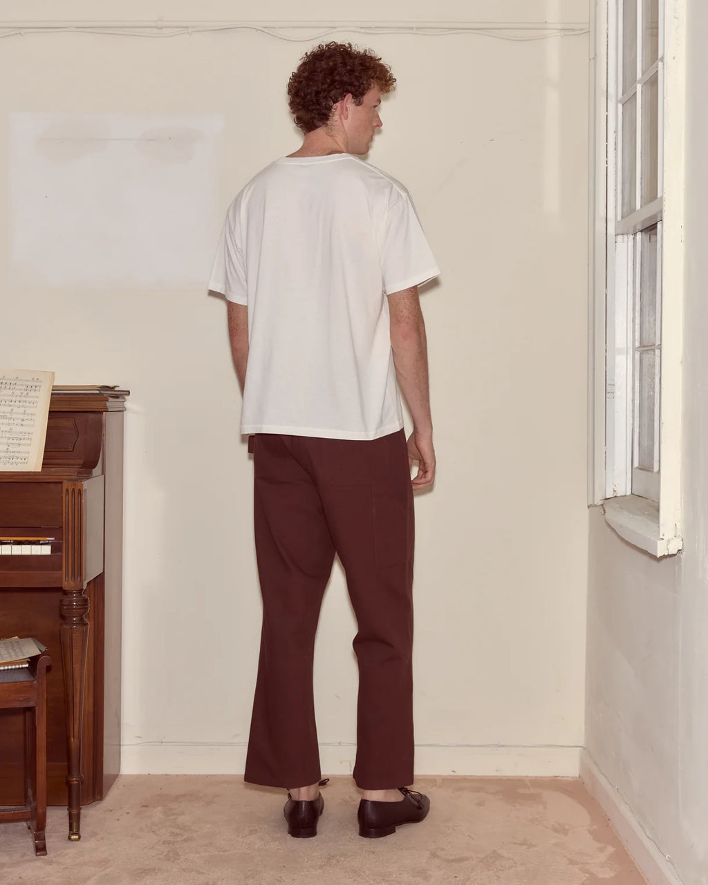 Person wearing a white t-shirt and brown pants standing in a room with a piano and window.