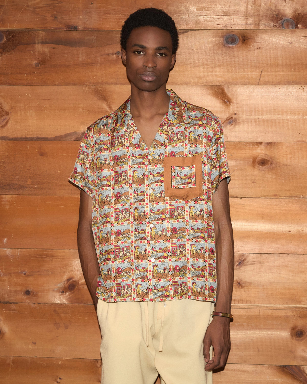 Model wearing Bode Cowboy Comic Short Sleeve Shirt in multicolor silk with a repeating cowboy comic print and front patch pocket.