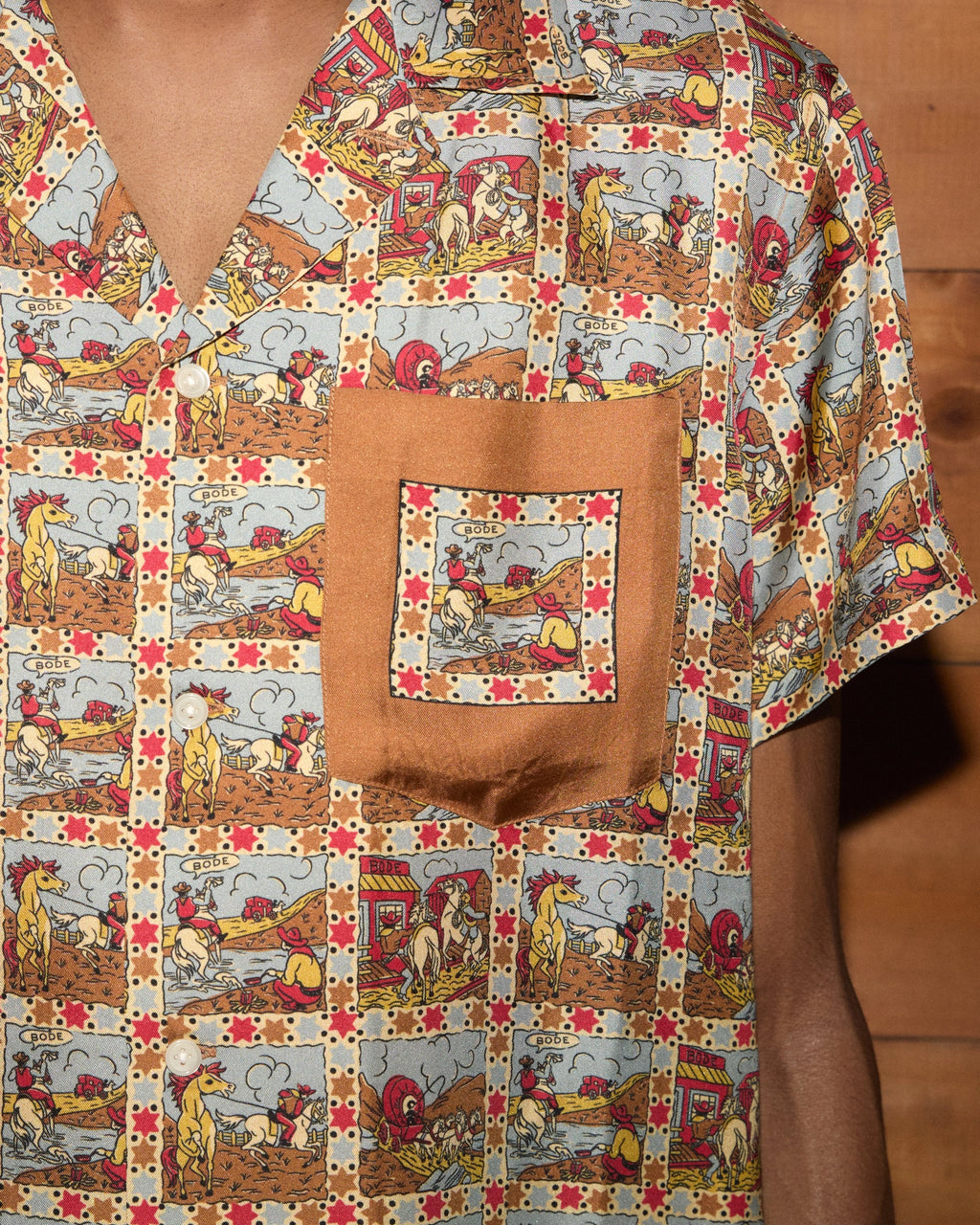 Model wearing Bode Cowboy Comic Short Sleeve Shirt in multicolor silk with a repeating cowboy comic print and front patch pocket.