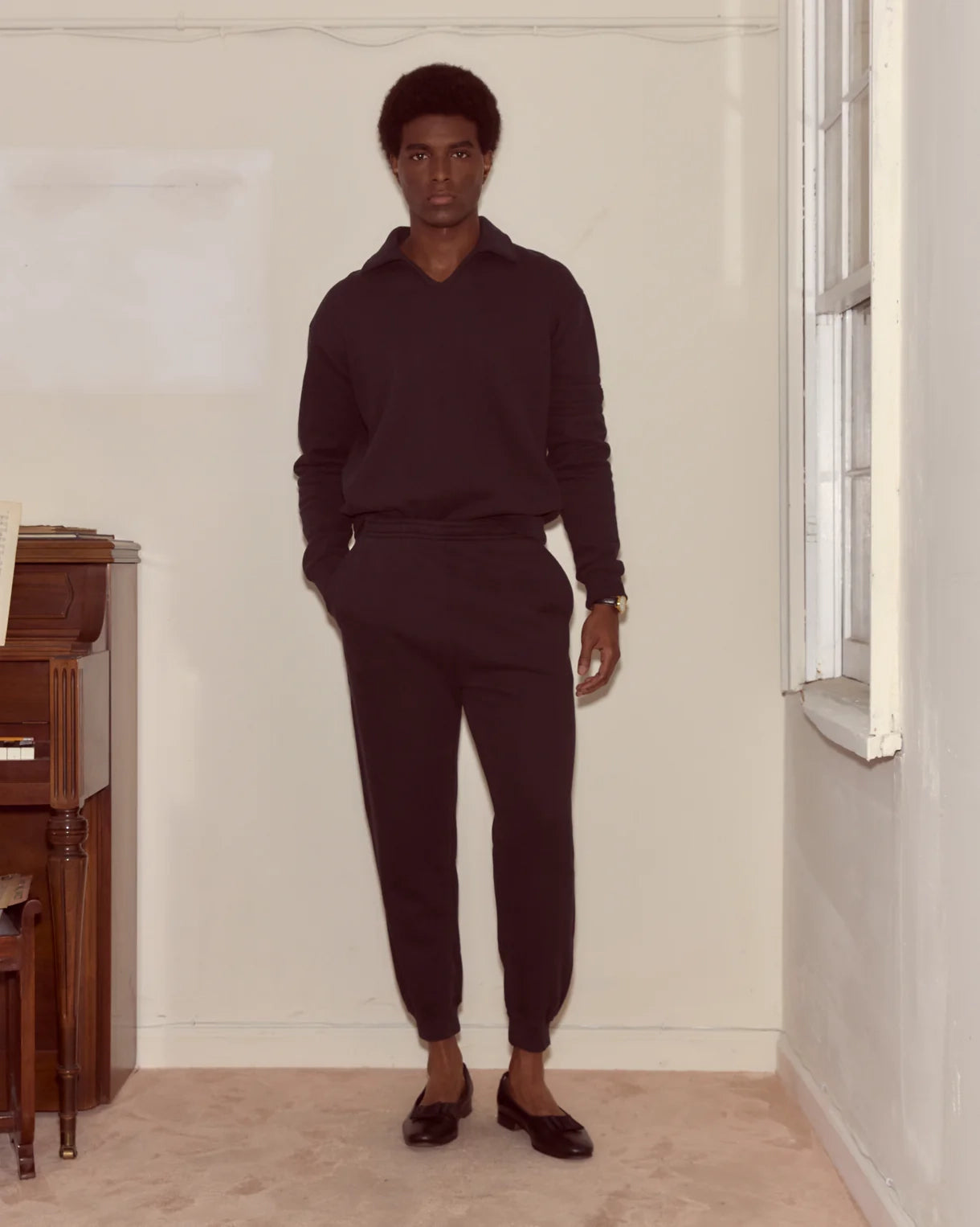 Man wearing a dark sweater and pants standing in a room with white walls and wooden furniture.