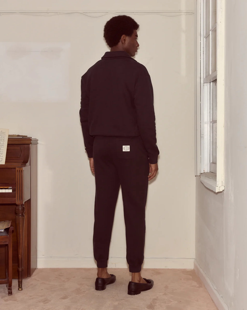 Person wearing a dark outfit standing in a room with a piano and window.
