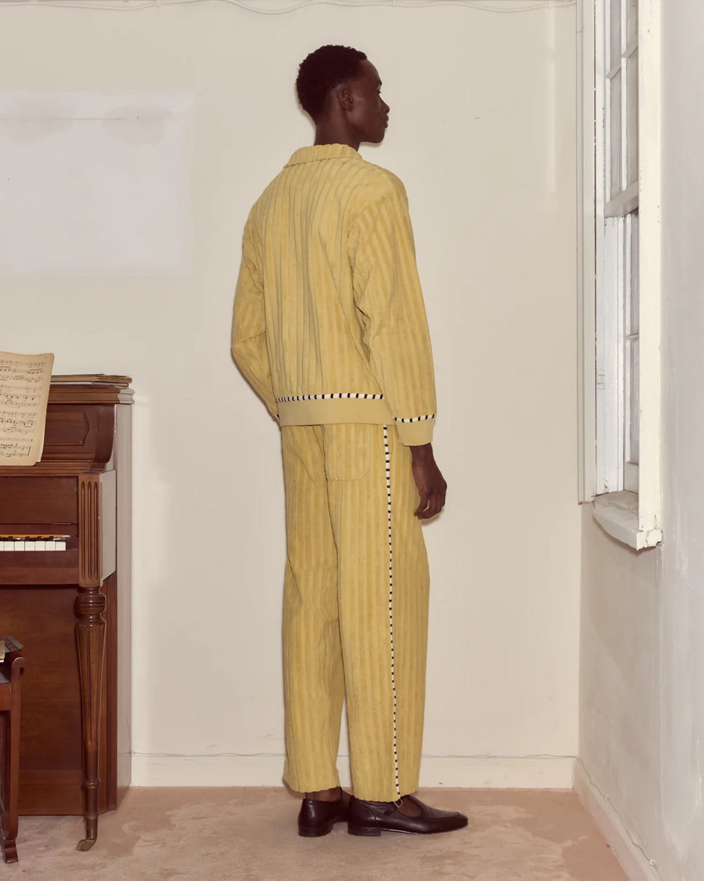 Person wearing a yellow outfit standing in a room with a piano and window.