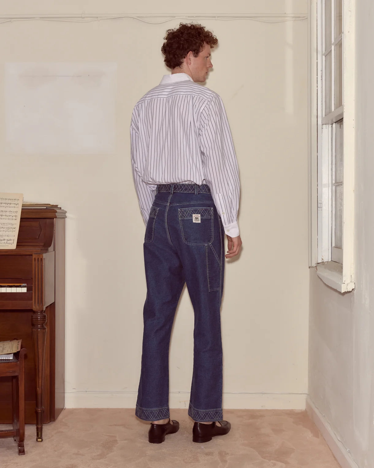 Person wearing a striped shirt and blue jeans standing in a room with a piano.