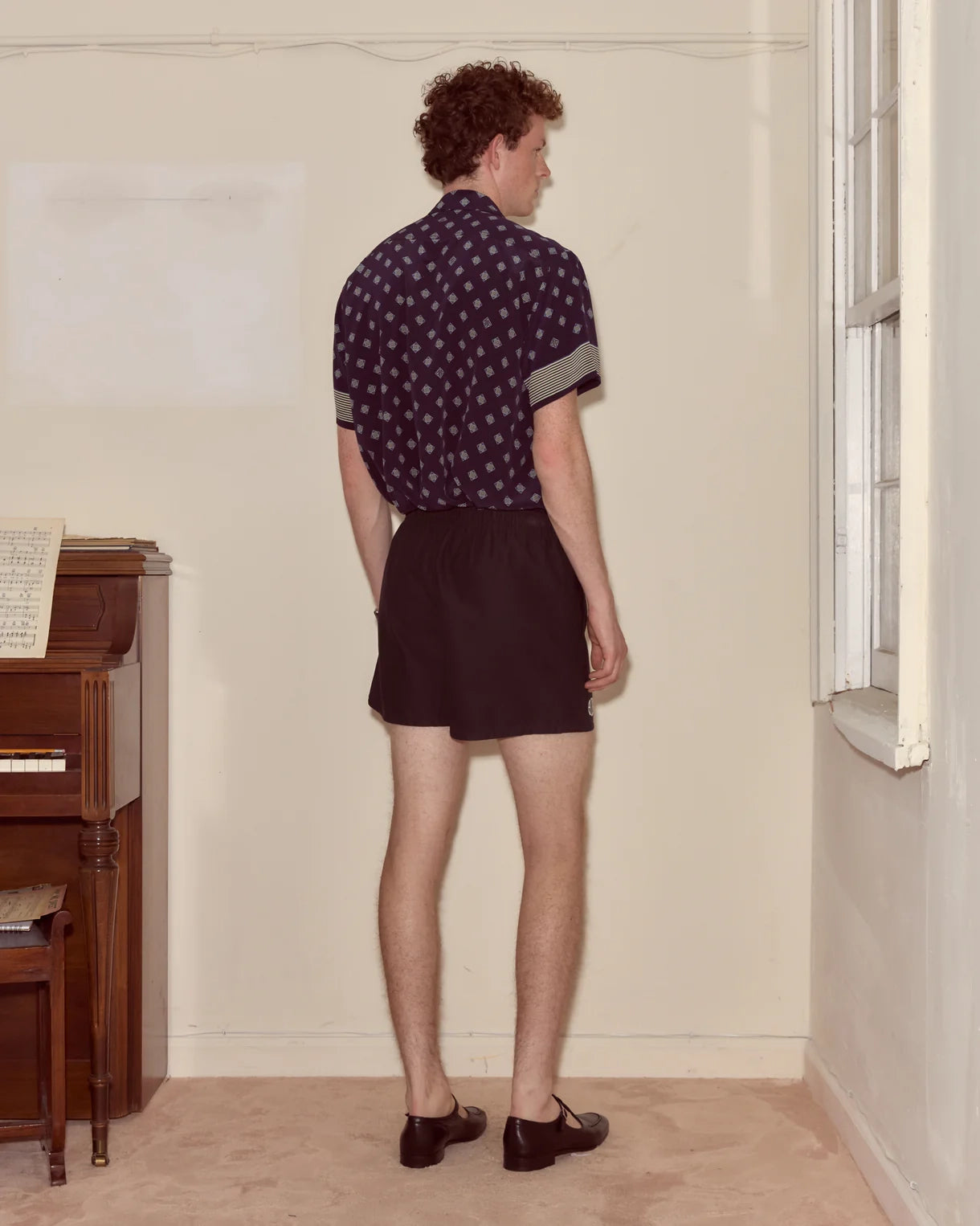 Person wearing a patterned shirt and shorts standing in a room with a piano.