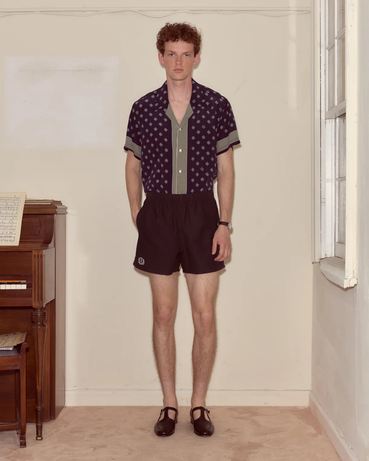 Man wearing a patterned shirt and dark shorts standing in a room with a piano.