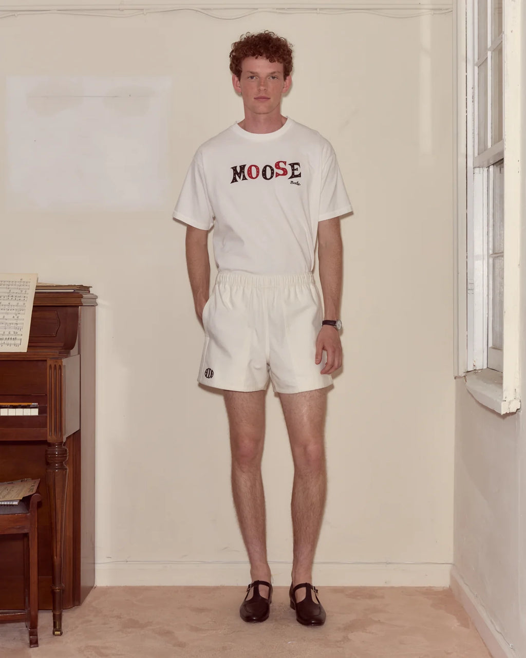 Man wearing a white t-shirt with 'MOOSE' print and white shorts in a room with a piano.