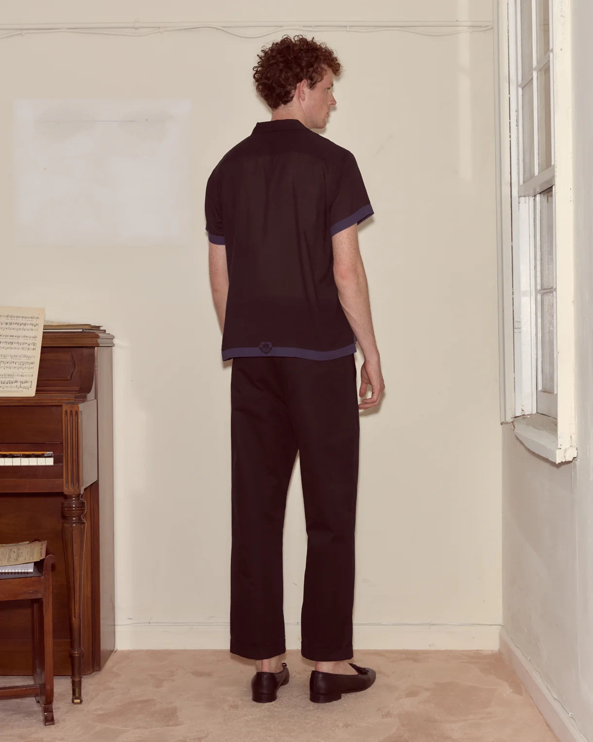 Person wearing a dark outfit standing in a room with a piano and window.