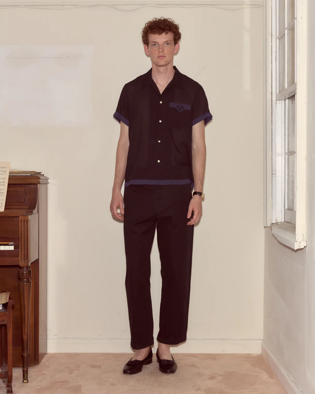 Man wearing a dark shirt and pants standing in a room with a window and wooden furniture.