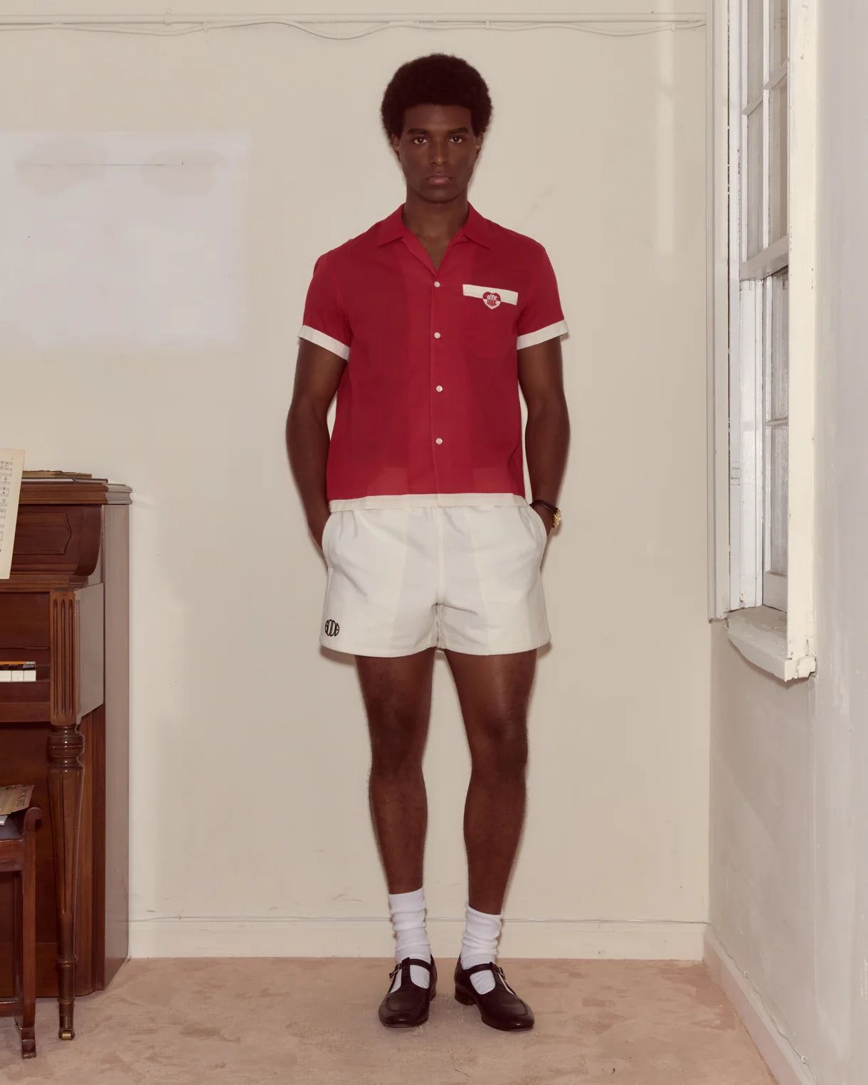 Man wearing a red shirt and white shorts standing in a room with a piano.