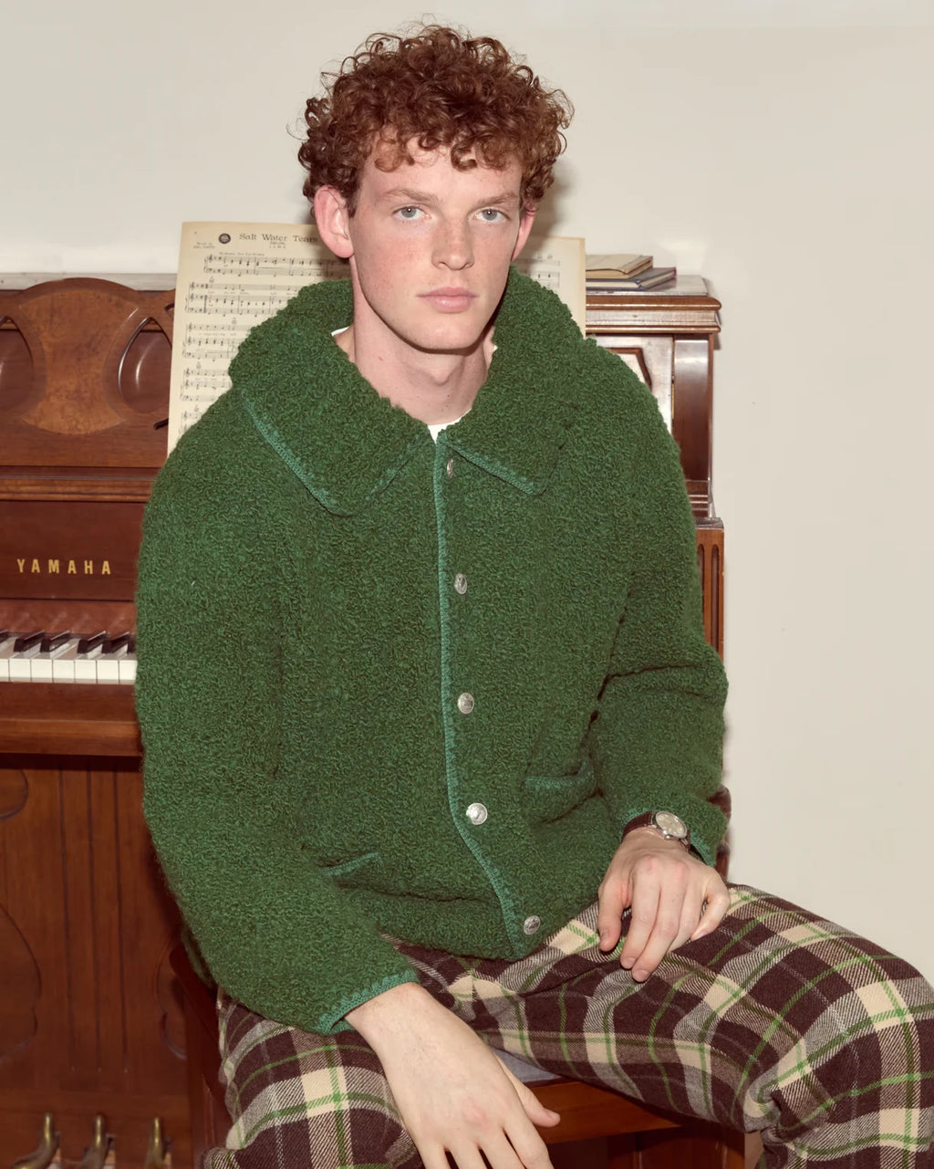 Person wearing a green jacket and plaid pants sitting in front of a piano.