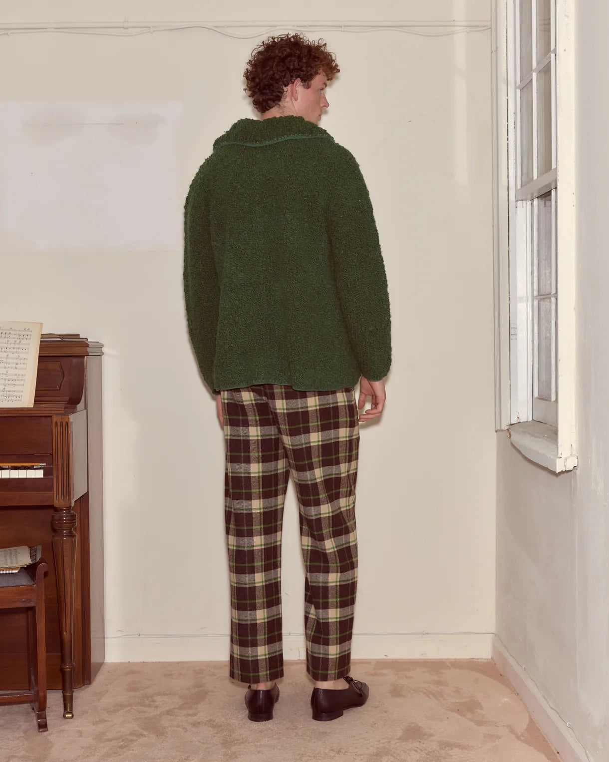 Person wearing a green sweater and plaid pants standing in a room with a piano.