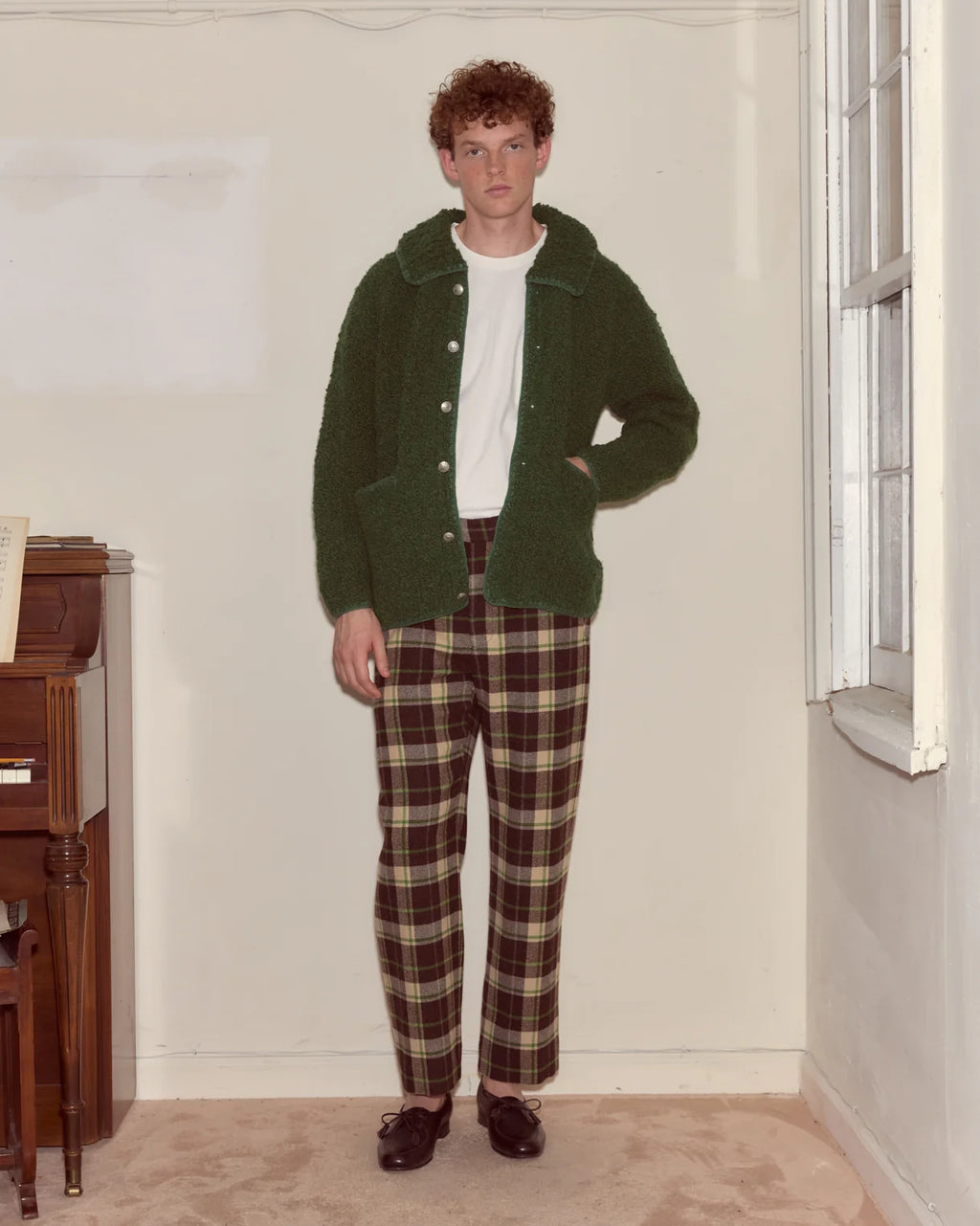 Person wearing a green cardigan and plaid pants standing in a room with a window and wooden furniture.