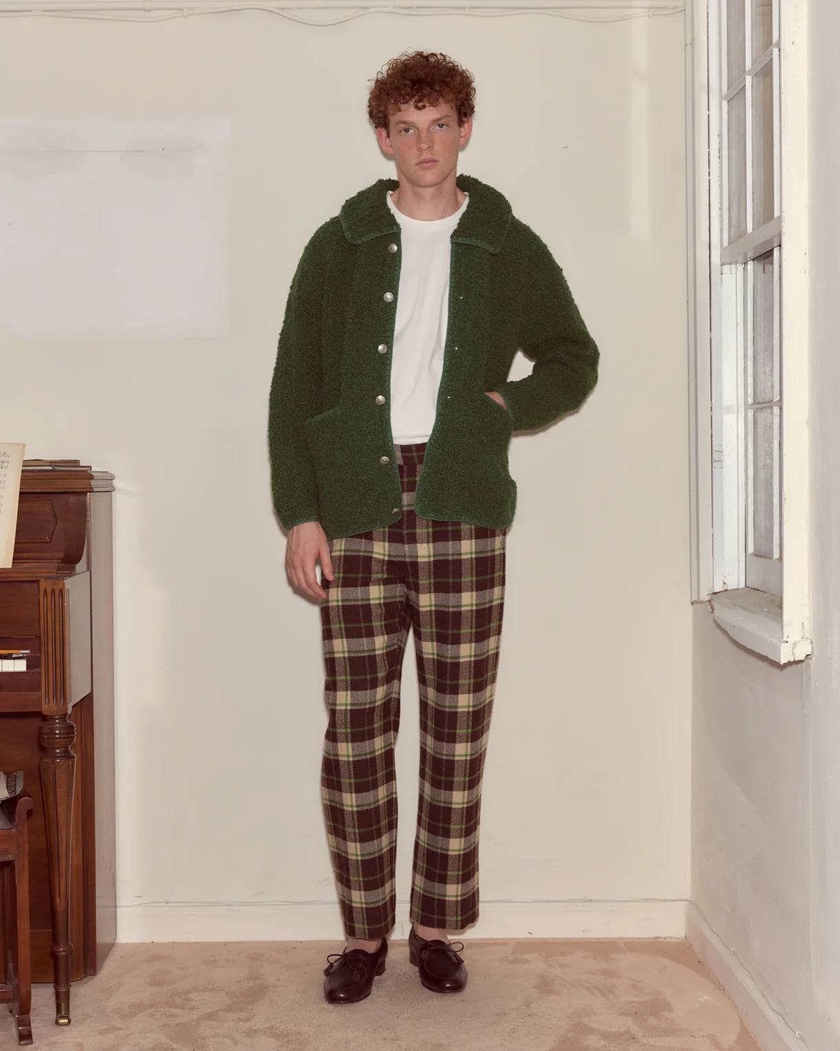Person wearing a green cardigan and plaid pants standing in a room with a window and wooden furniture.