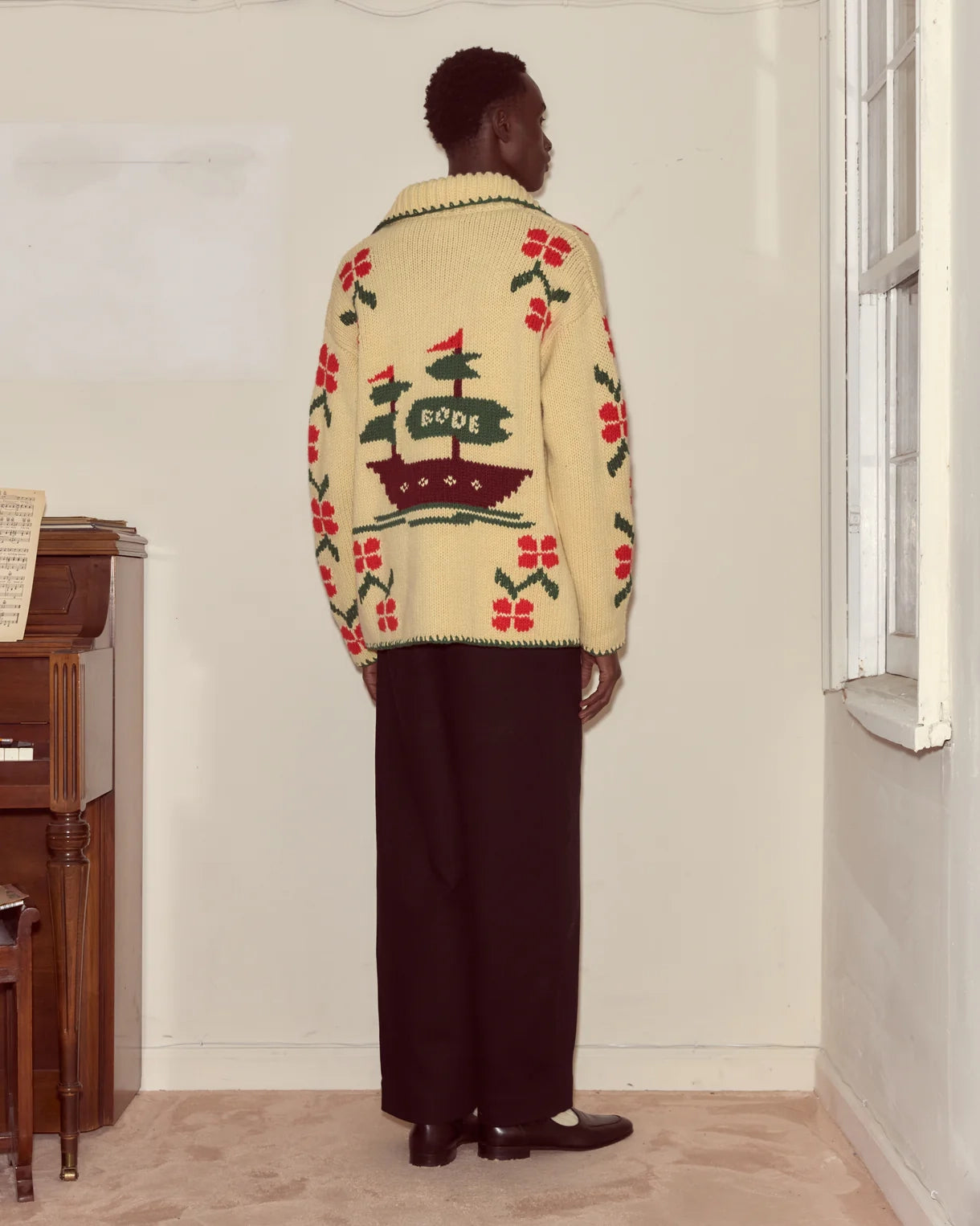Person wearing a patterned sweater in a room with a window and wooden furniture.