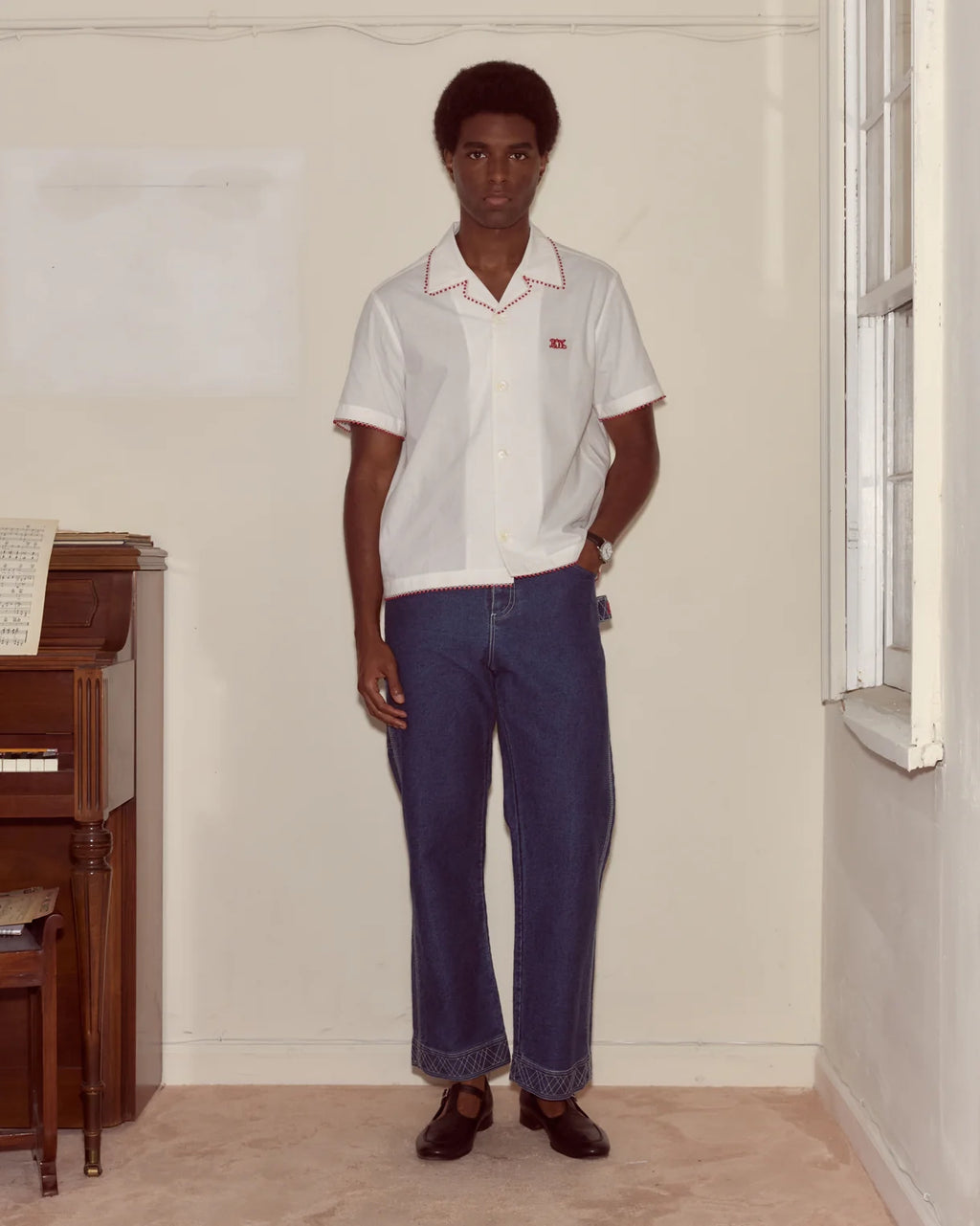 Person wearing a white polo shirt and blue jeans standing in a room with a piano.