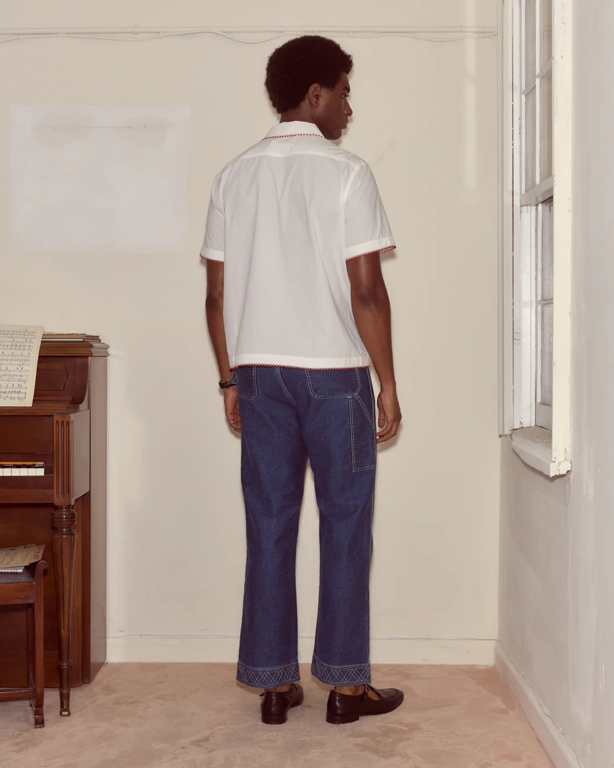 Person wearing a white polo shirt and blue jeans standing in a room with a piano.