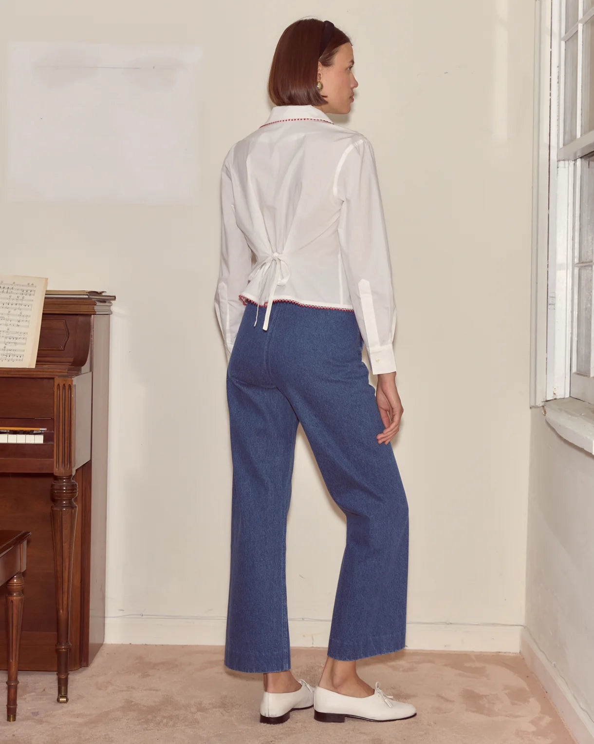 Woman wearing a white blouse and blue pants standing in a room with a piano and window.