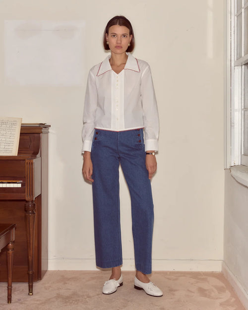 Woman wearing a white blouse with red trim and blue pants standing in a room with a piano.