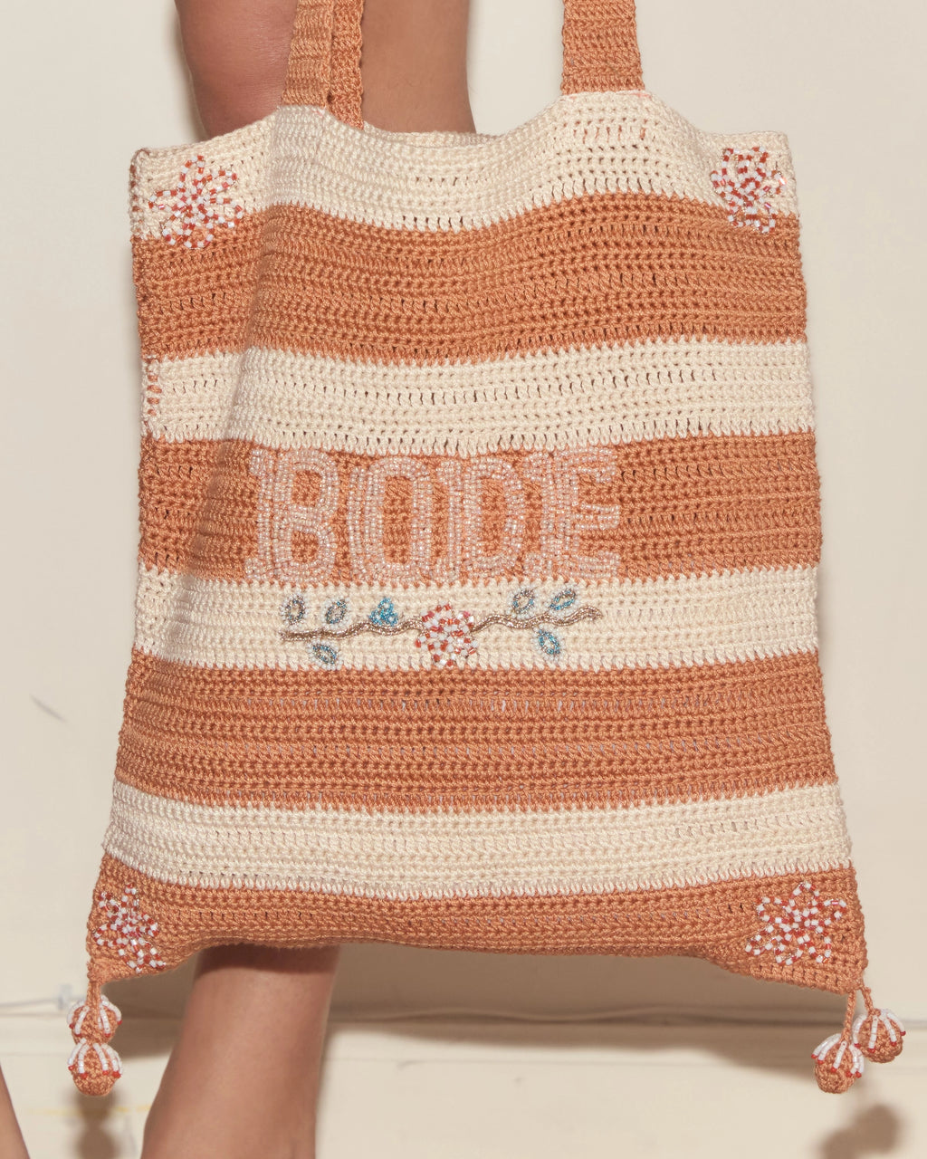 Model holding orange and cream Bode crochet tote bag with striped panels, floral beadwork and Bode lettering.