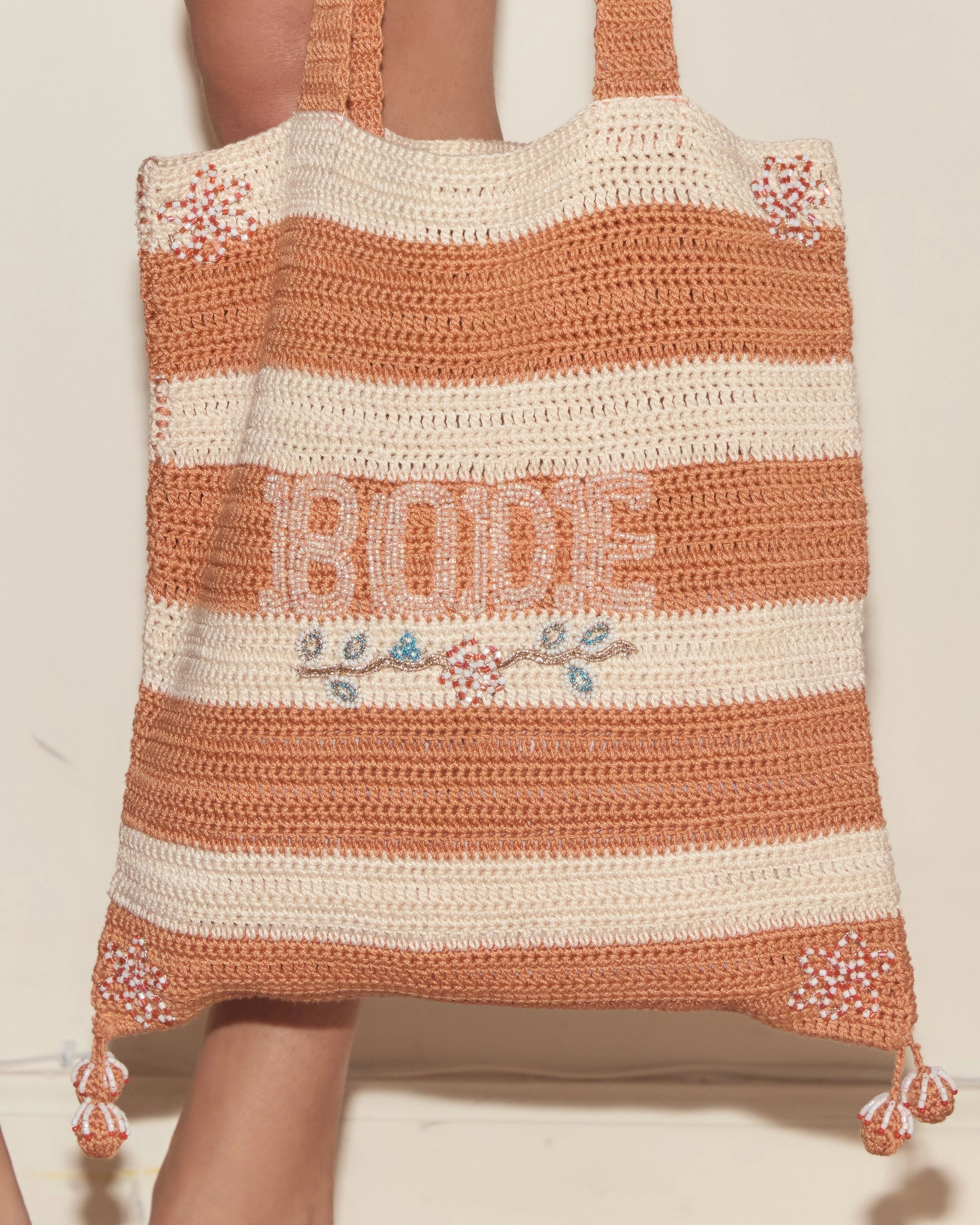 Model holding orange and cream Bode crochet tote bag with striped panels, floral beadwork and Bode lettering.