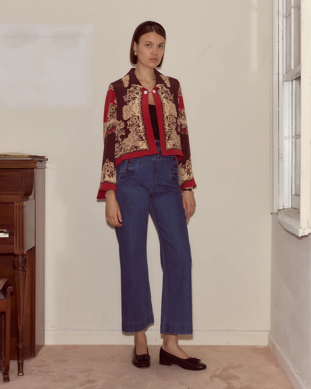 Woman wearing a patterned jacket and blue jeans standing in a room with a piano.