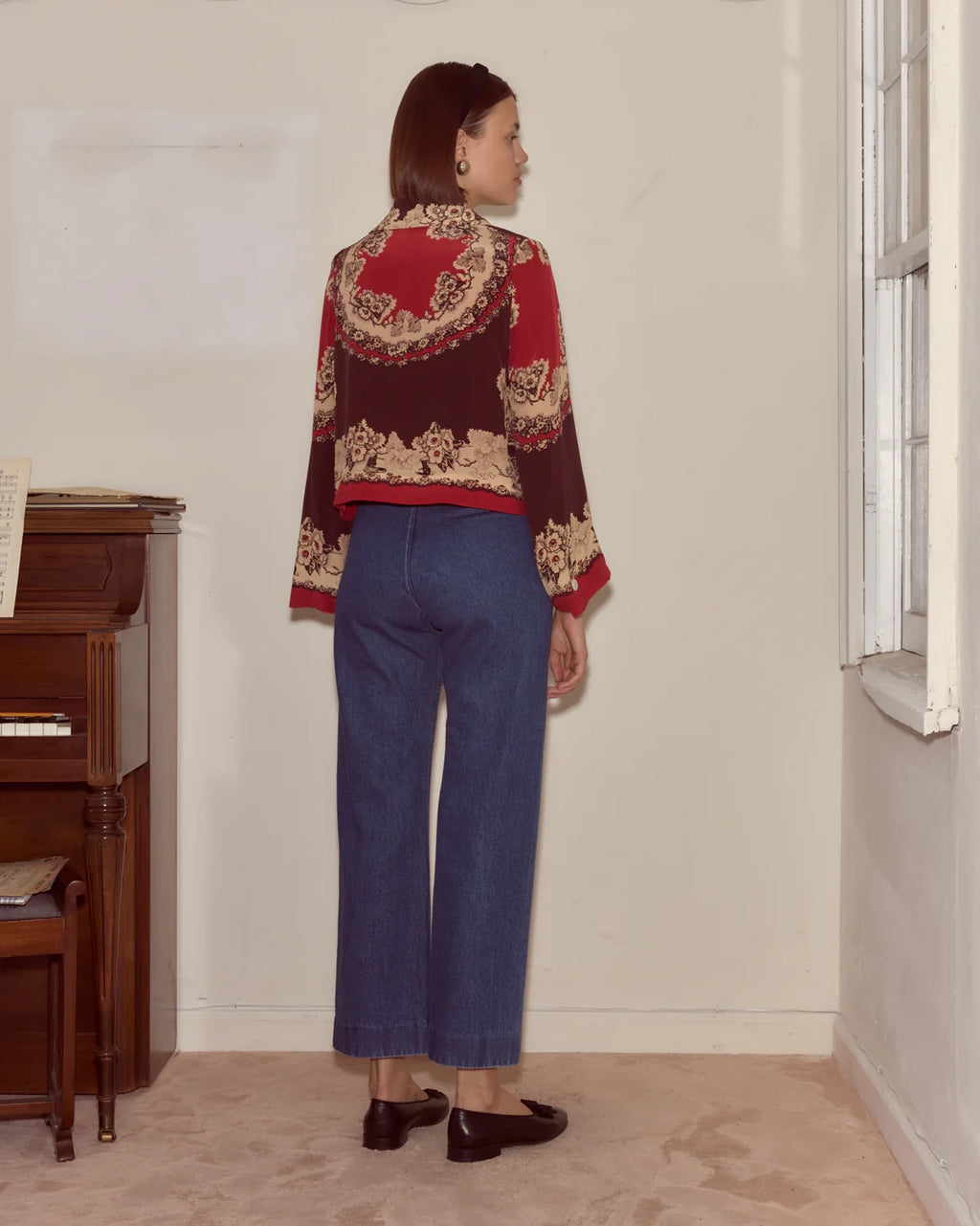 Person wearing a patterned red and gold sweater and blue jeans standing in a room with a piano.