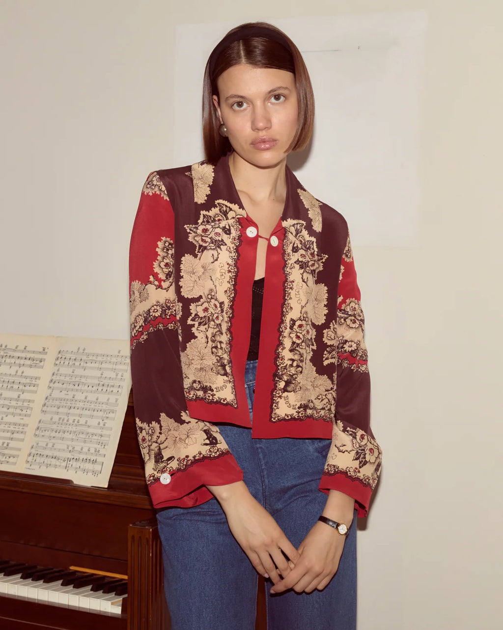 Woman wearing a red and beige patterned jacket standing next to a piano with sheet music.