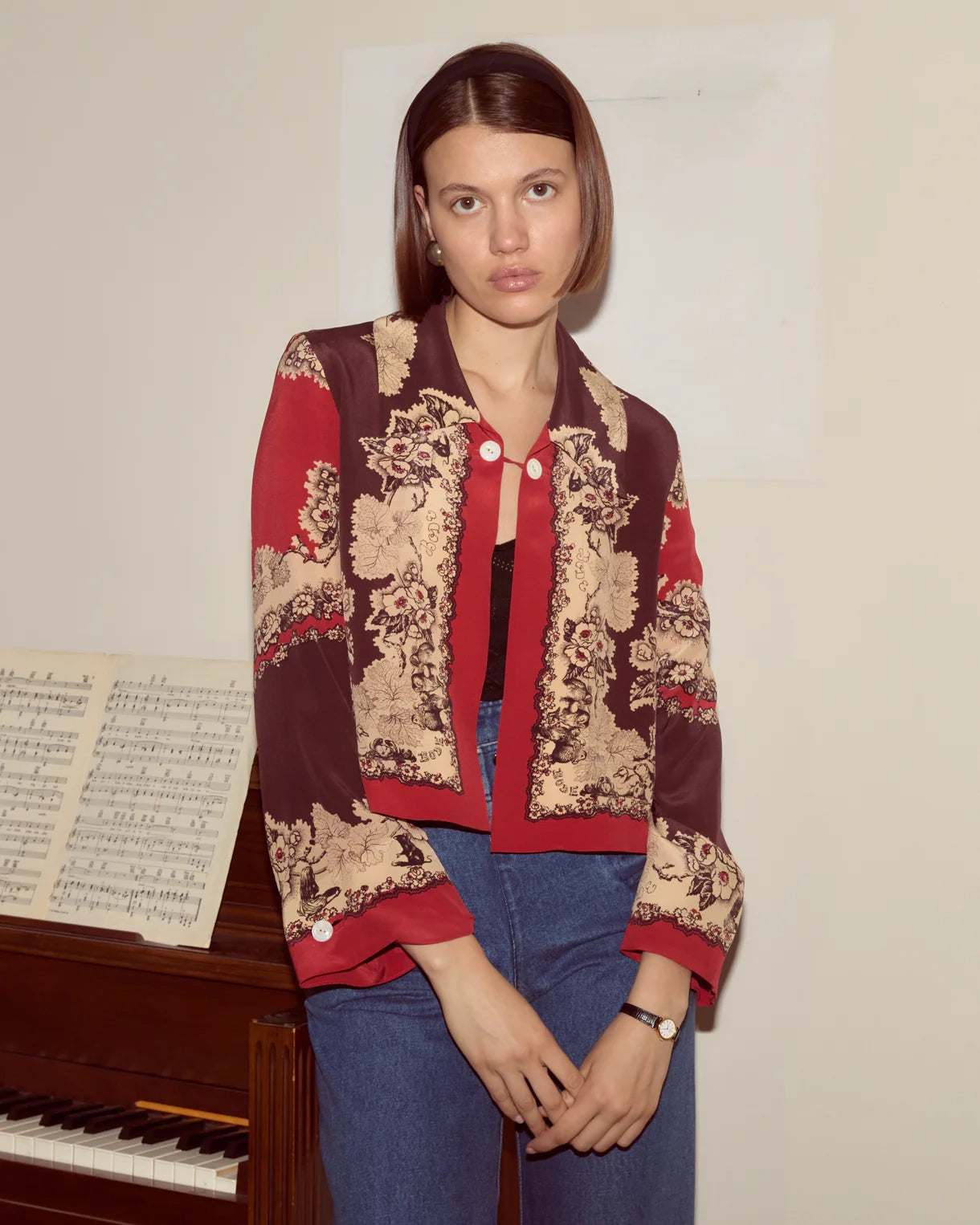 Woman wearing a red and beige patterned jacket standing next to a piano with sheet music.
