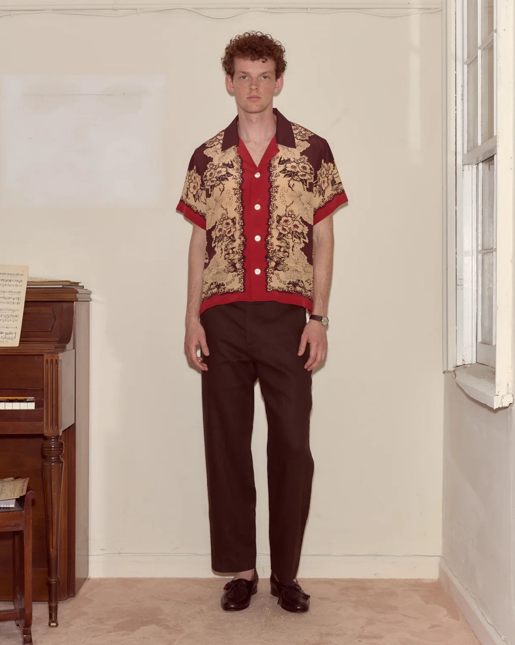 Man wearing a patterned shirt and dark pants standing in a room with a piano.