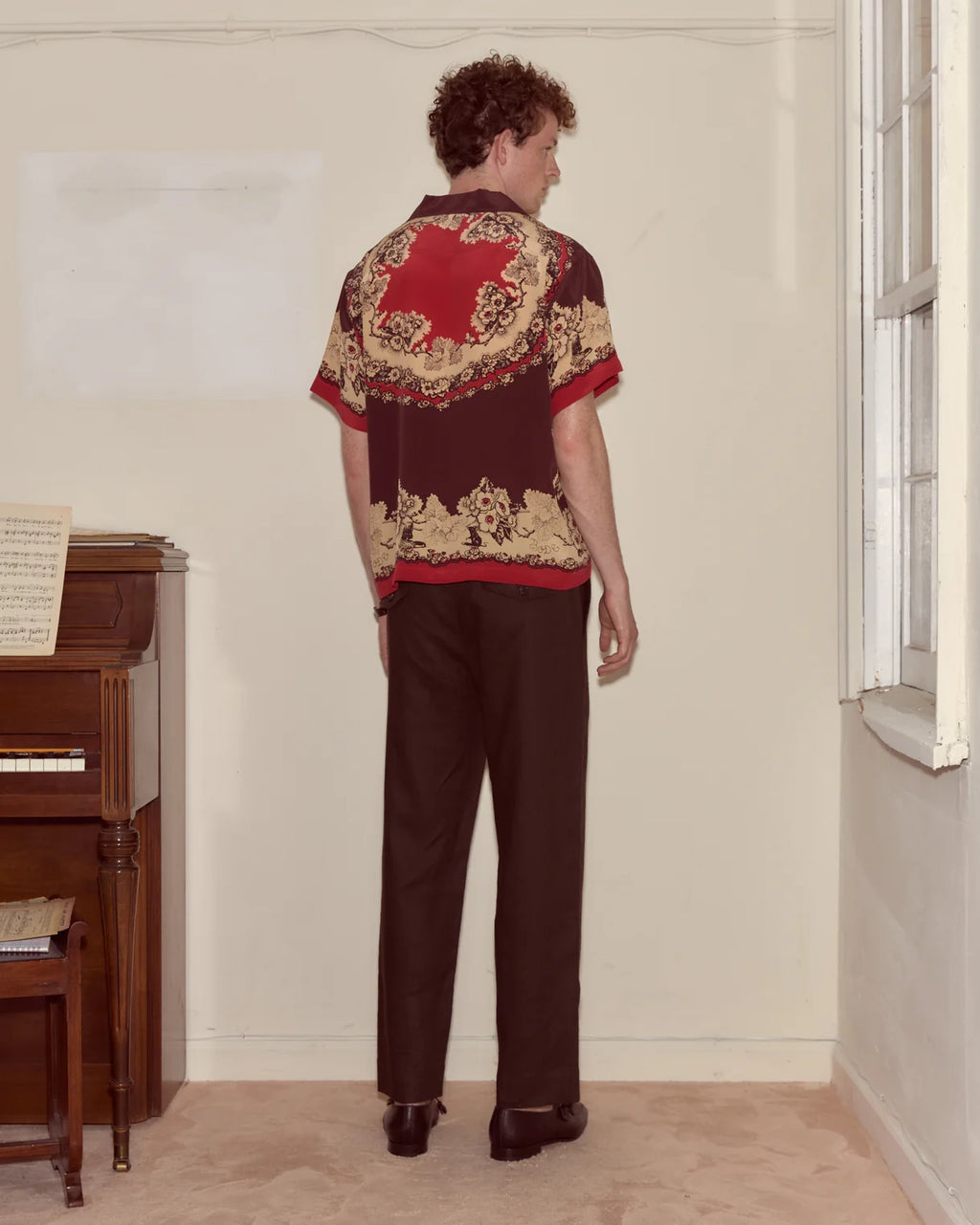 Person wearing a patterned shirt and pants standing in a room with a piano.