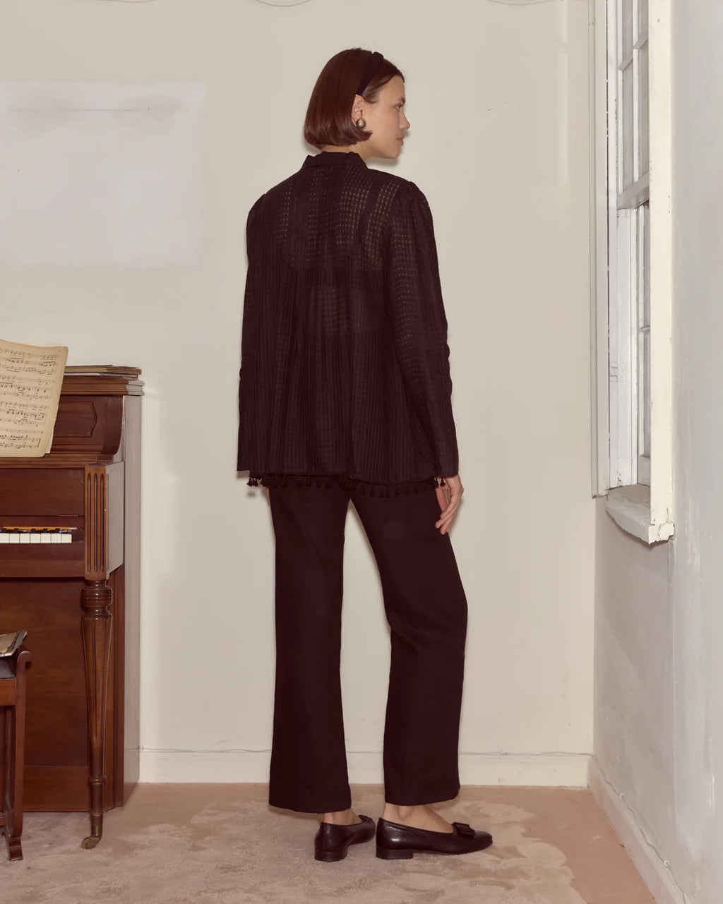 Woman in a dark outfit standing in a room with a piano and window.