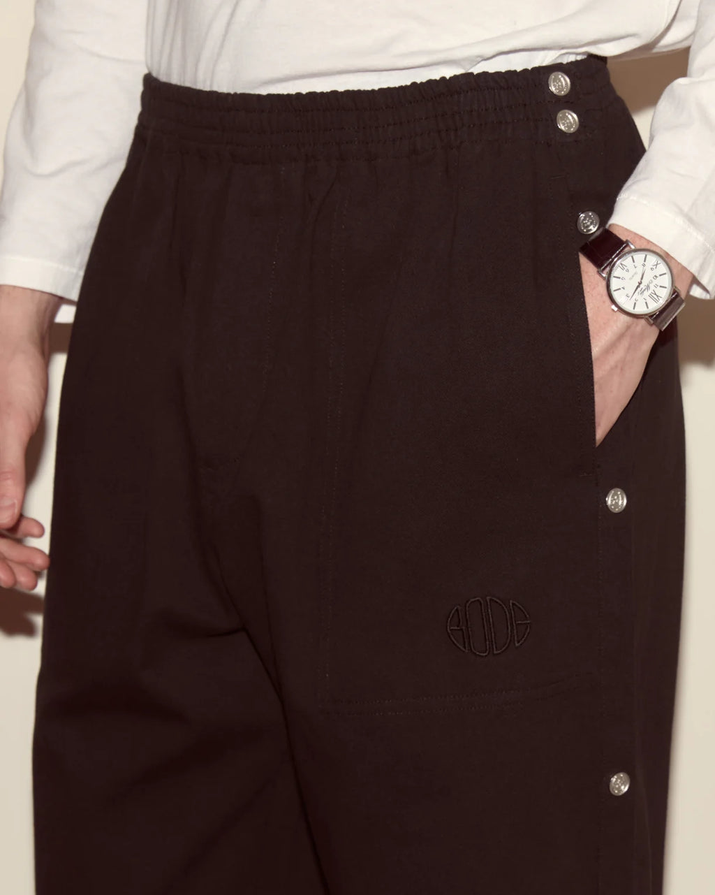 Person wearing black pants with button details and a white shirt, holding a watch.