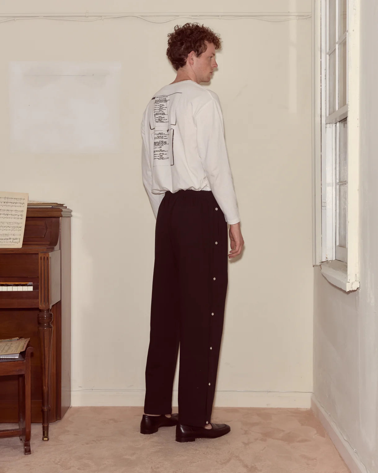 Person wearing a white shirt with text and dark pants standing in a room with a piano.