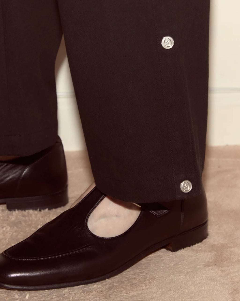 Close-up of dark brown leather shoes and pants with button details on a neutral background