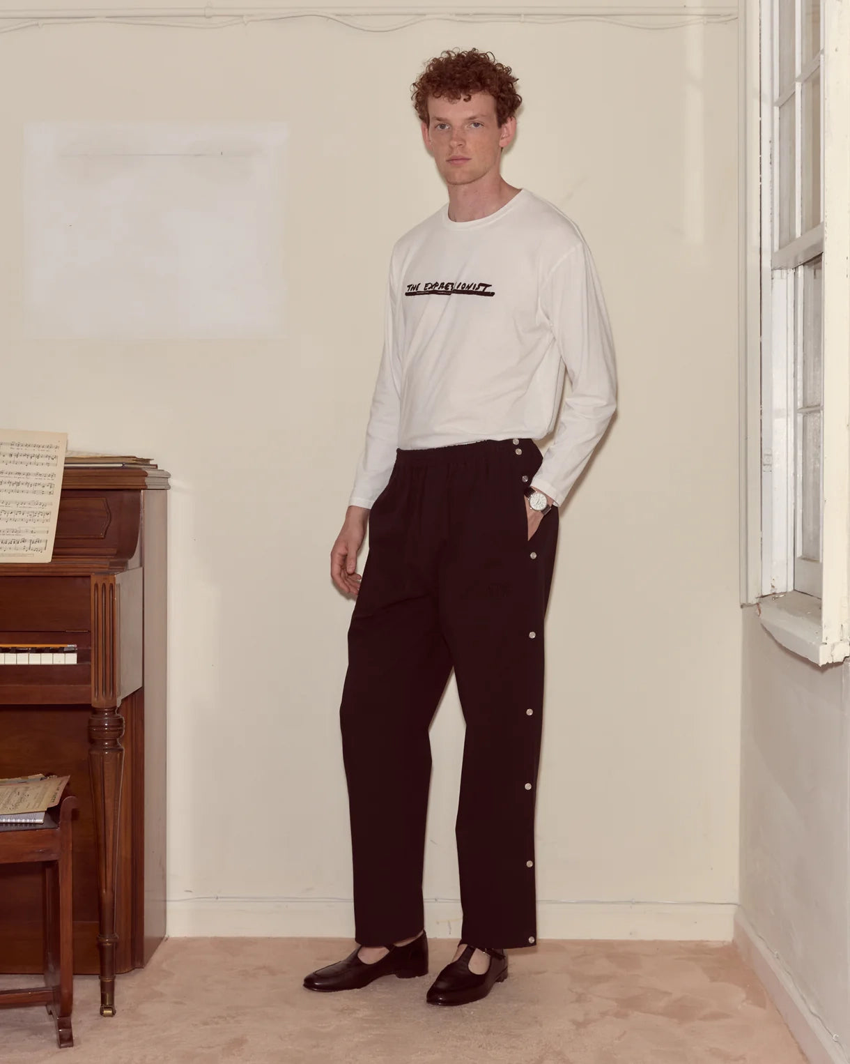 Man wearing a white long-sleeve shirt and dark pants standing in a room with a piano.