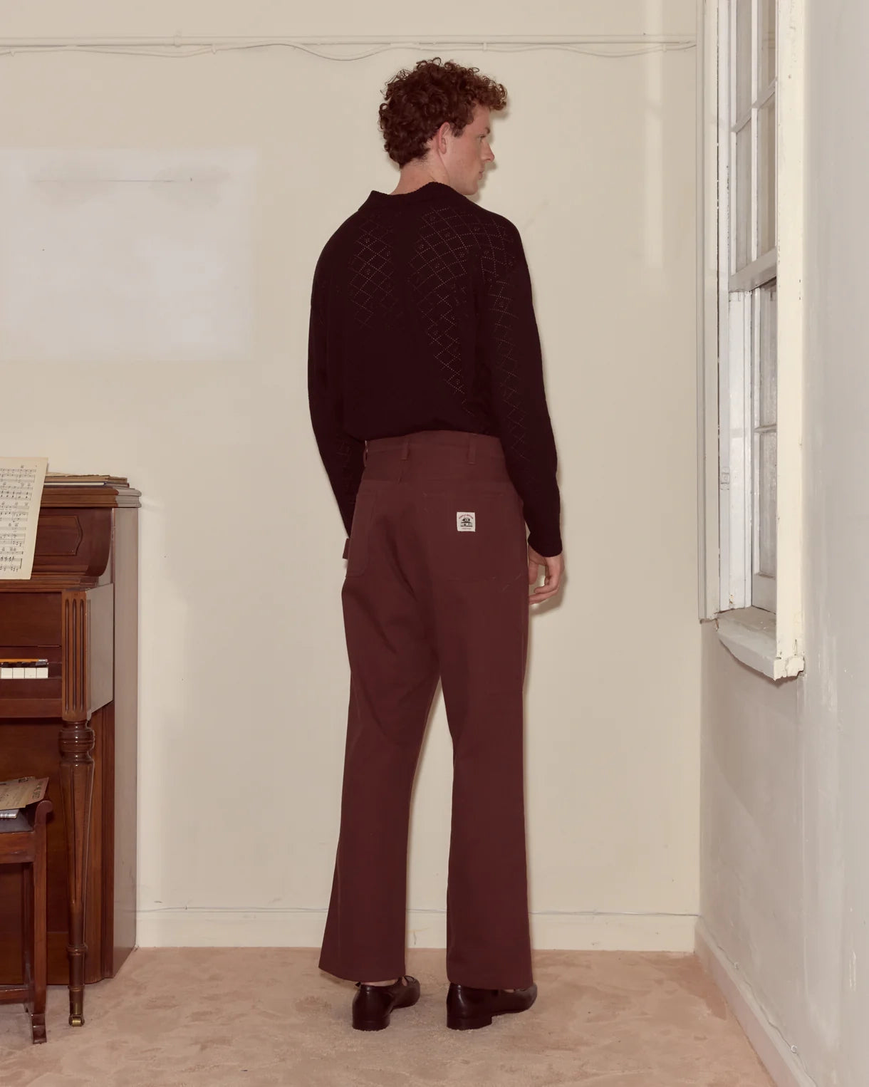 Person wearing a dark sweater and maroon pants standing in a room with a window and wooden furniture.